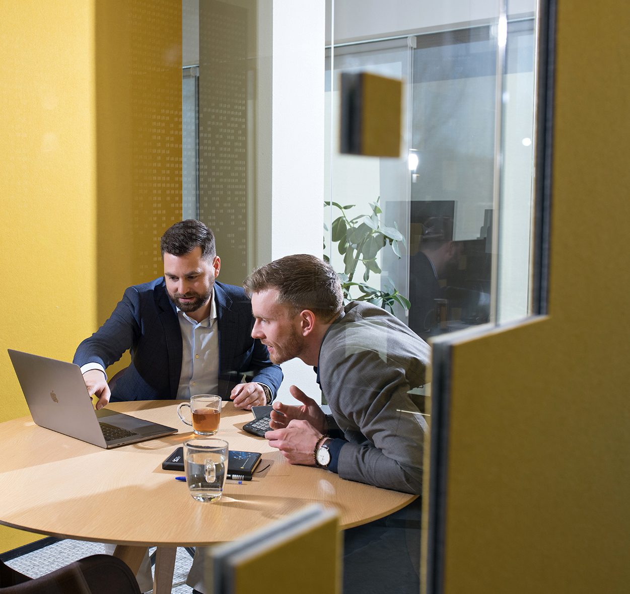 Two men are seated at a round wooden table in a modern office space with yellow walls. They are both focused on a laptop screen, with one man pointing at the screen while the other leans forward attentively. The table also has a glass of water, a notebook, a pen, and a cup of tea. In the background, there is a glass wall, allowing a view into another part of the office with a potted plant visible. The setting is professional and collaborative, suggesting a focused discussion or work session.