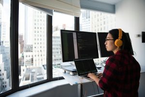 A software developer wearing headphones, working on multiple monitors with code, in a high-rise office, symbolizing the focus on green software engineering and energy-efficient coding practices.