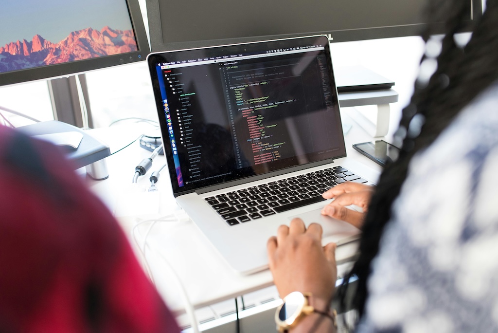 A person is working on a laptop, writing or reviewing code displayed on the screen. The coding environment features a dark theme with colorful syntax highlighting. The workspace includes additional monitors, one of which shows a scenic mountain landscape. Various office supplies and electronic devices are present on the desk, creating a professional and tech-savvy atmosphere. The scene suggests a software development or programming task.