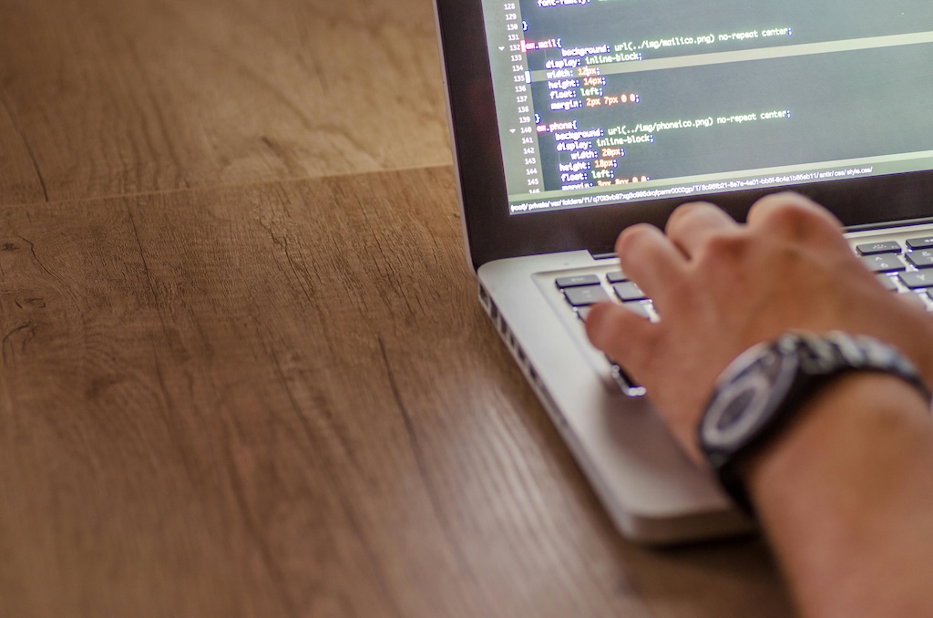 A person working on a laptop, with their hand on the keyboard. The screen displays lines of code in a text editor, suggesting programming or coding activity. The background features a wooden table, creating a simple and focused workspace environment. The person is wearing a wristwatch, adding a personal touch to the scene.